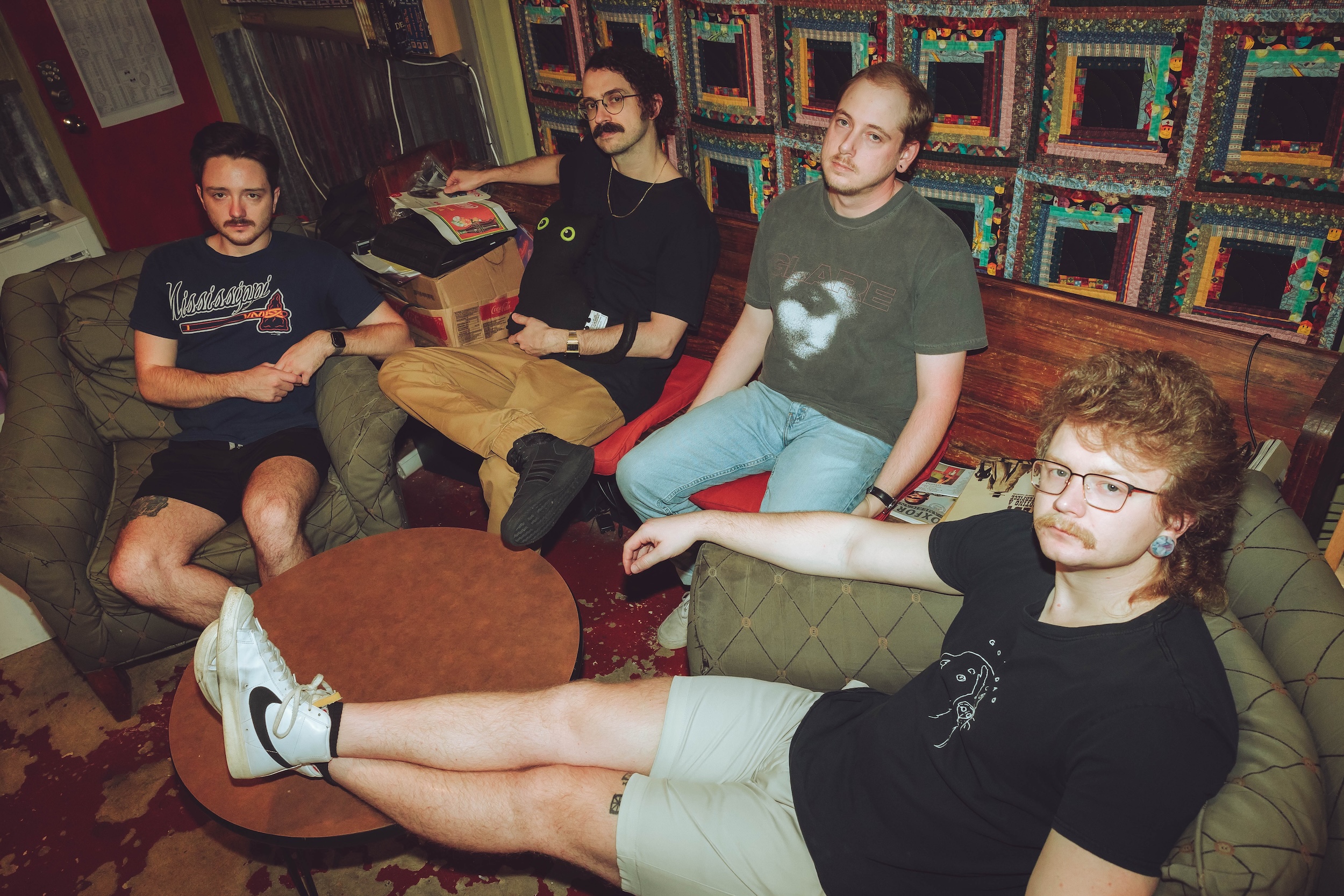 Inside Voices band promo image A promotional photo of the band Inside Voices, featuring four members seated in a cozy, eclectic room. On the left, a man in a navy Mississippi-themed T-shirt and black shorts sits on a green armchair. Next to him, another member wearing a black shirt, tan pants, and glasses holds a black stuffed toy with green eyes. On the right, a man in a faded gray graphic T-shirt and jeans sits on a red bench, while another member with a mullet hairstyle, glasses, a mustache, and a black shirt with a minimalist design lounges with his legs stretched out, wearing white Nike sneakers. The room is decorated with a colorful patchwork quilt on the wall, wooden furniture, and a patterned carpet.