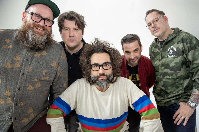 Five members of the band Motion City Soundtrack pose together in a brightly lit studio with a white backdrop. Front and center, lead singer Justin Pierre leans forward wearing a cream-colored sweatshirt with colorful stripes, his curly hair tousled and glasses on. The other four bandmates stand behind him, smiling or looking toward the camera—one in a bird-patterned shirt and cap, another in a black button-up, one in a maroon jacket, and one in a camo windbreaker with "The Player Agency" logo. The mood is casual and friendly. Photo by Ben Pier.