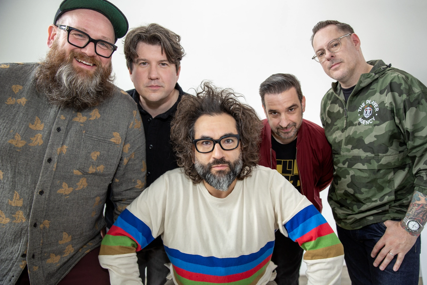 Five members of the band Motion City Soundtrack pose together in a brightly lit studio with a white backdrop. Front and center, lead singer Justin Pierre leans forward wearing a cream-colored sweatshirt with colorful stripes, his curly hair tousled and glasses on. The other four bandmates stand behind him, smiling or looking toward the camera—one in a bird-patterned shirt and cap, another in a black button-up, one in a maroon jacket, and one in a camo windbreaker with "The Player Agency" logo. The mood is casual and friendly. Photo by Ben Pier.