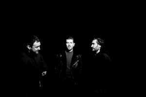 Twin Atlantic band photo: three members photographed in near-darkness, lit from above, wearing dark clothing, in a moody black and white portrait